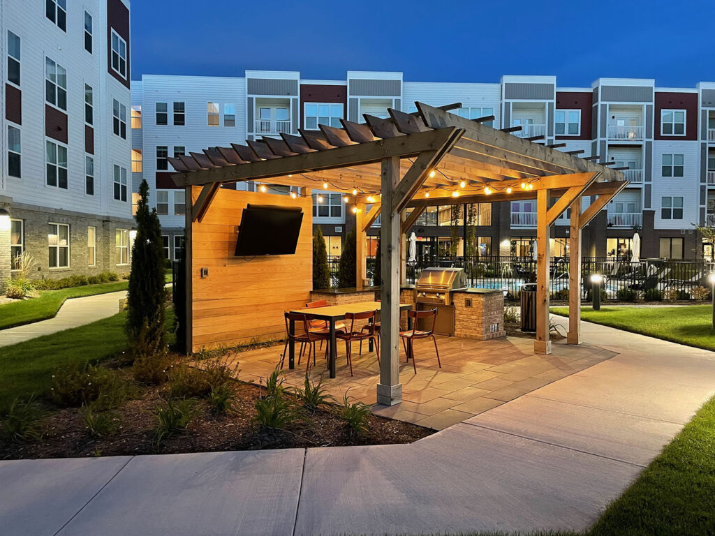 Outdoor kitchen with grill and black granite countertop. An overhead pergola features string lights for nighttime ambiance. Shiplap wall with mounted outdoor TV provides entertainment for guests sitting at dining table